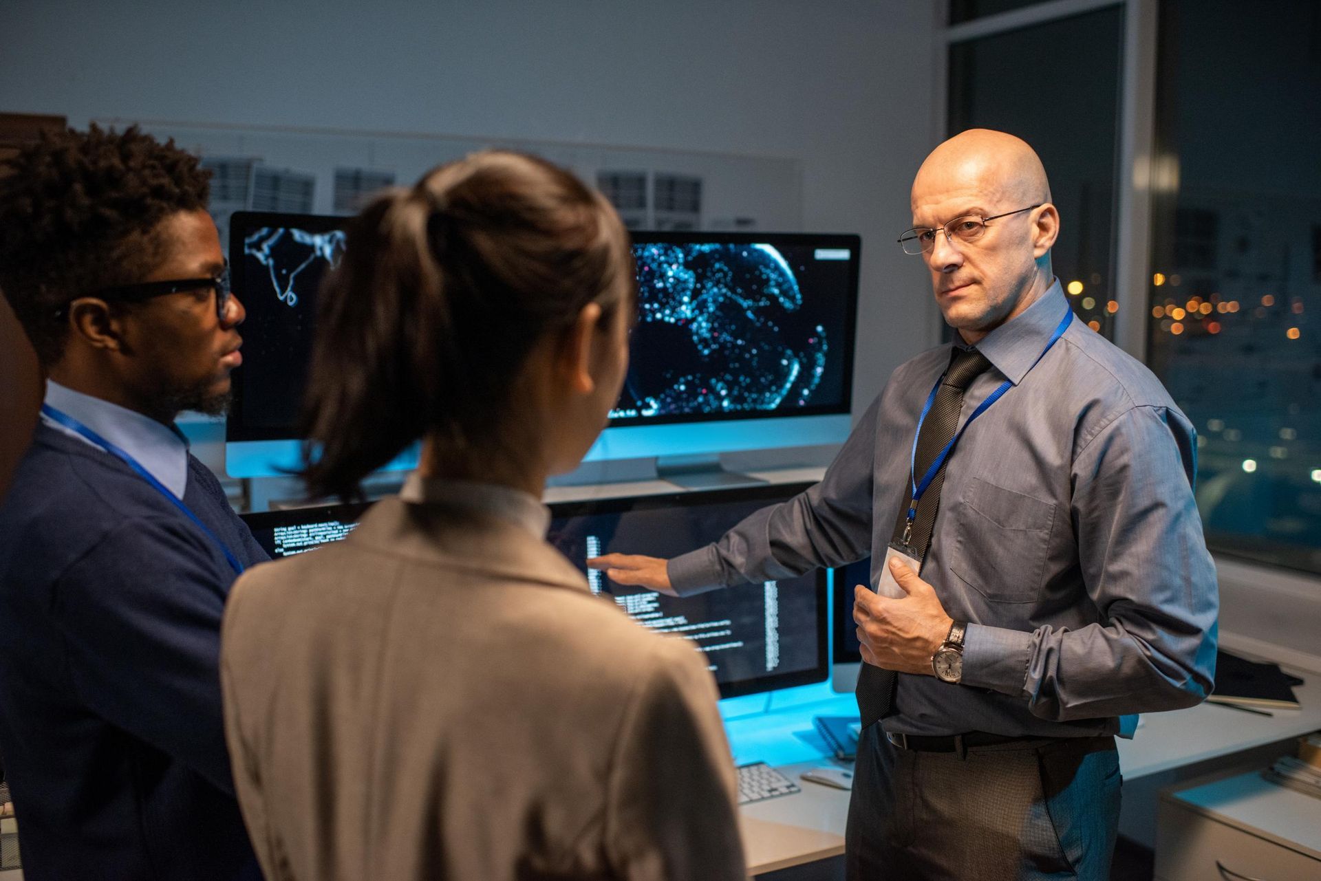 Three people in a dark control room look at computer screens. A man points to a screen as he speaks.