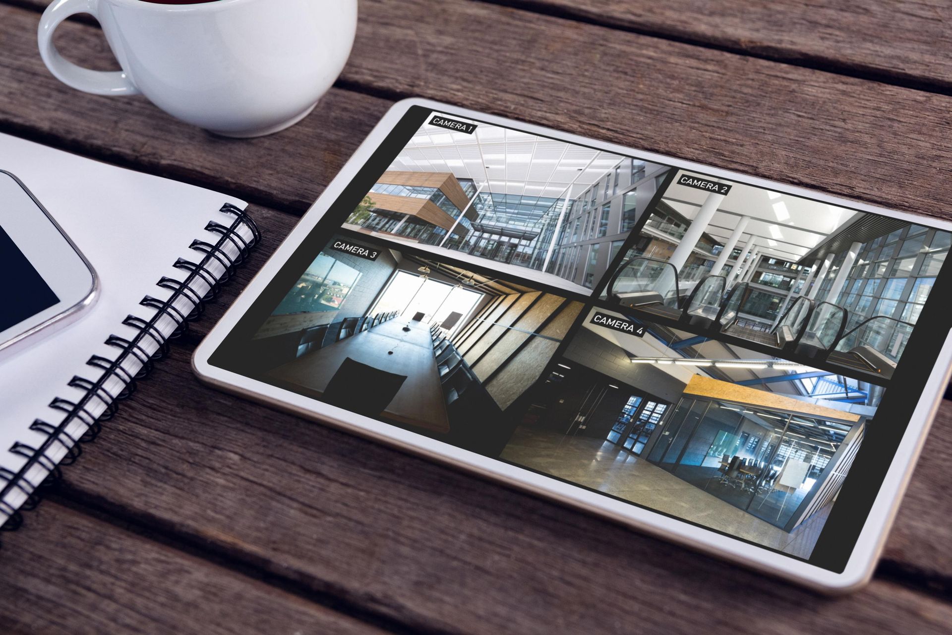 Tablet displaying security camera footage on a wooden table next to a coffee cup, notepad, and smartphone.