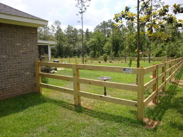 A wooden fence surrounds a lush green yard in front of a brick house.