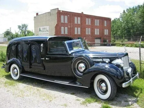 Black vintage hearse parked in front of a brick building. White-walled tires.
