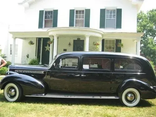 Black vintage hearse parked in front of a white house with green shutters.