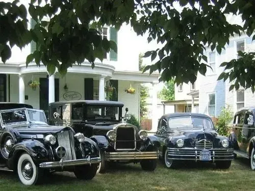 Classic cars parked on a lawn in front of a white house with a porch and trees.
