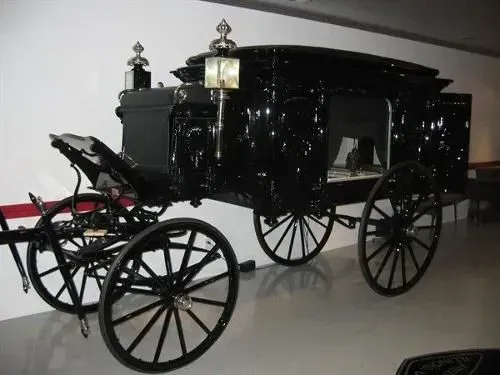 Black horse-drawn hearse in a museum. Glossy black finish, ornate details, four large spoked wheels.