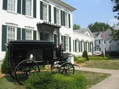 A black horse-drawn hearse in front of a white building with dark green shutters.