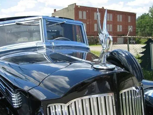 Black vintage car with chrome hood ornament in front of a brick building.