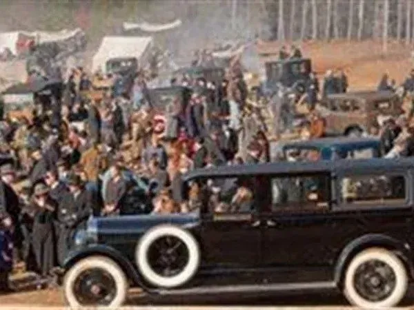 Crowd gathering around cars in an outdoor setting, likely a historical event with many people and tents.