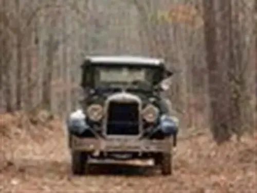Vintage black car driving on a dirt road in a wooded area.