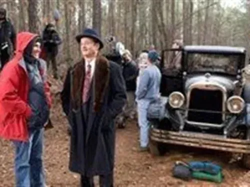 Man in fedora and coat, vintage car on a film set outdoors.