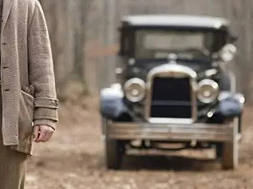 Man in beige jacket stands near vintage black car on a dirt road.