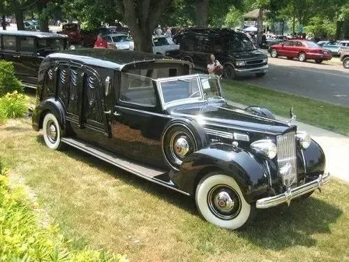 Black vintage hearse with white wall tires parked on grass.