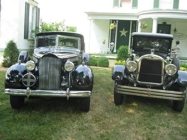 Two black vintage cars parked on a grassy lawn in front of a white house.