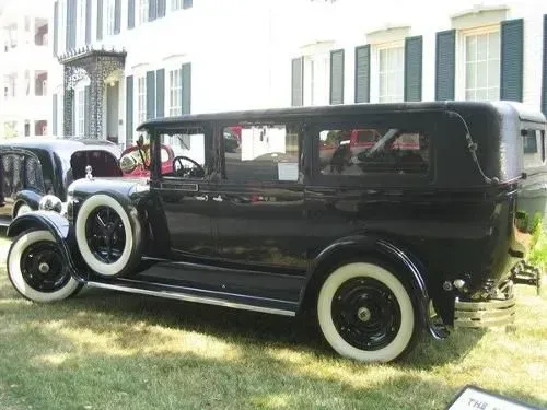 Black vintage car with white-walled tires parked on a lawn in front of a white building with columns.