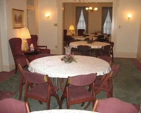 Round tables set in a room with chairs, a lamp, and an open doorway to another room with tables.
