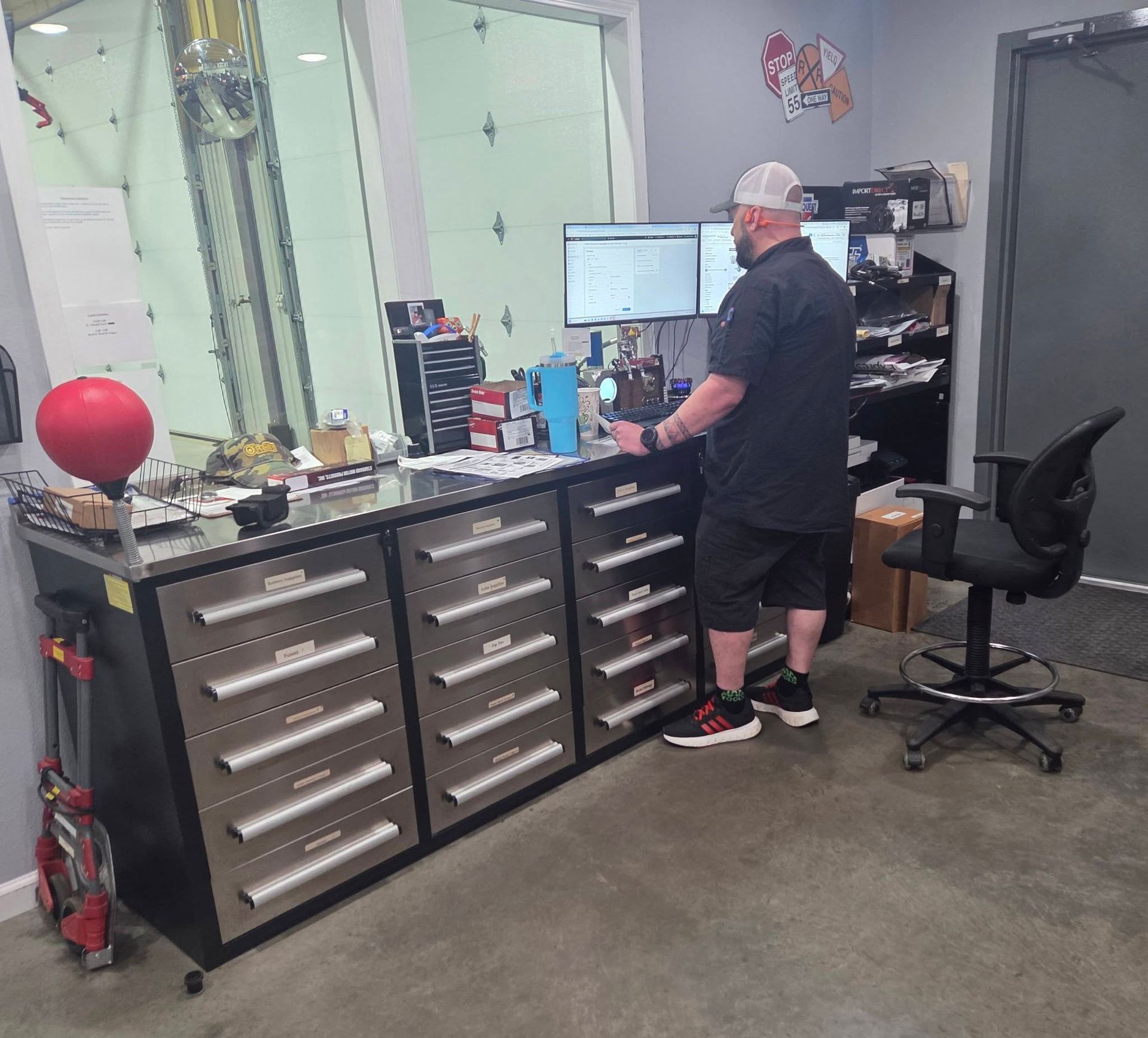 Person standing at a workbench in a garage workshop with drawers, tools, and computer monitors