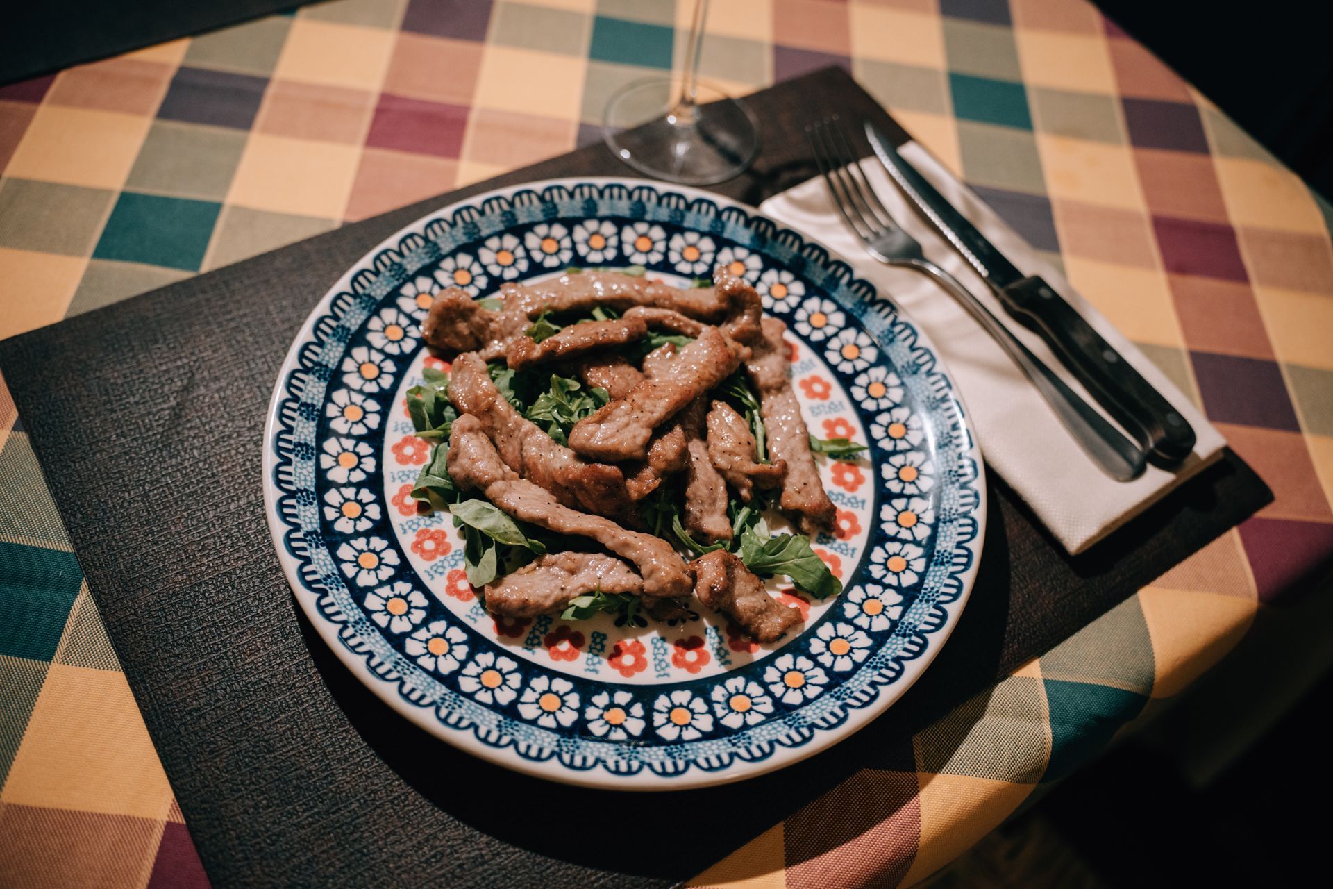Piatto di striscioline di carne su rucola, su un piatto decorato, su una tovaglia a quadretti con posate.