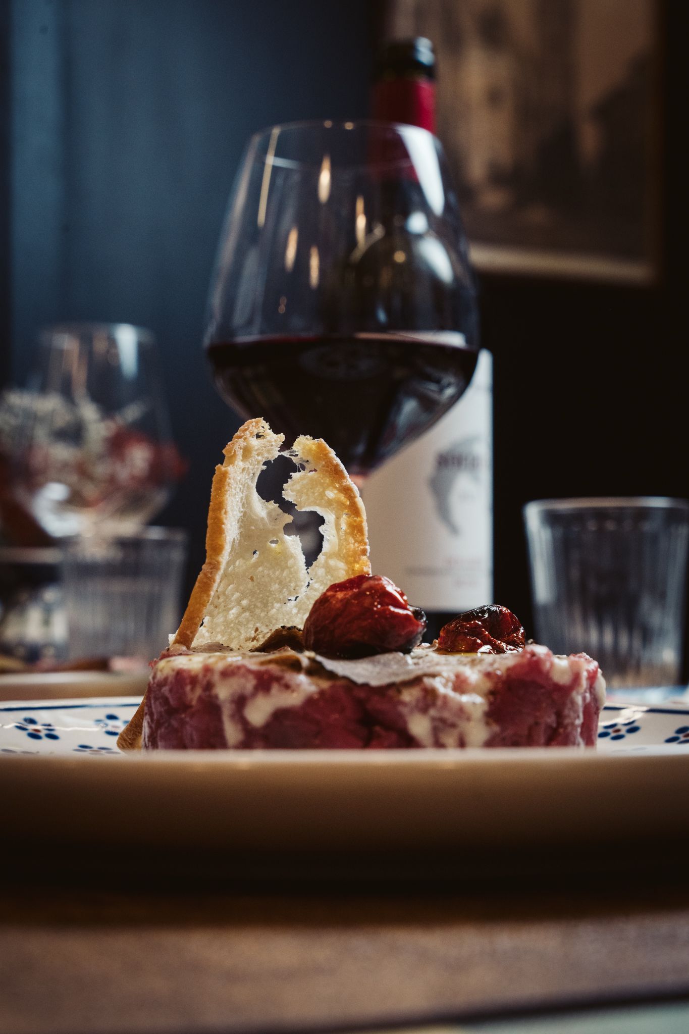 Piatto con condimenti, pane e un bicchiere di vino rosso su un tavolo con una bottiglia di vino sullo sfondo.