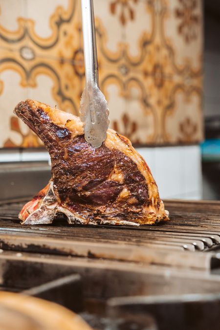 Una grande bistecca scottata viene girata sulla griglia con delle pinze. La griglia ha delle barre di metallo.