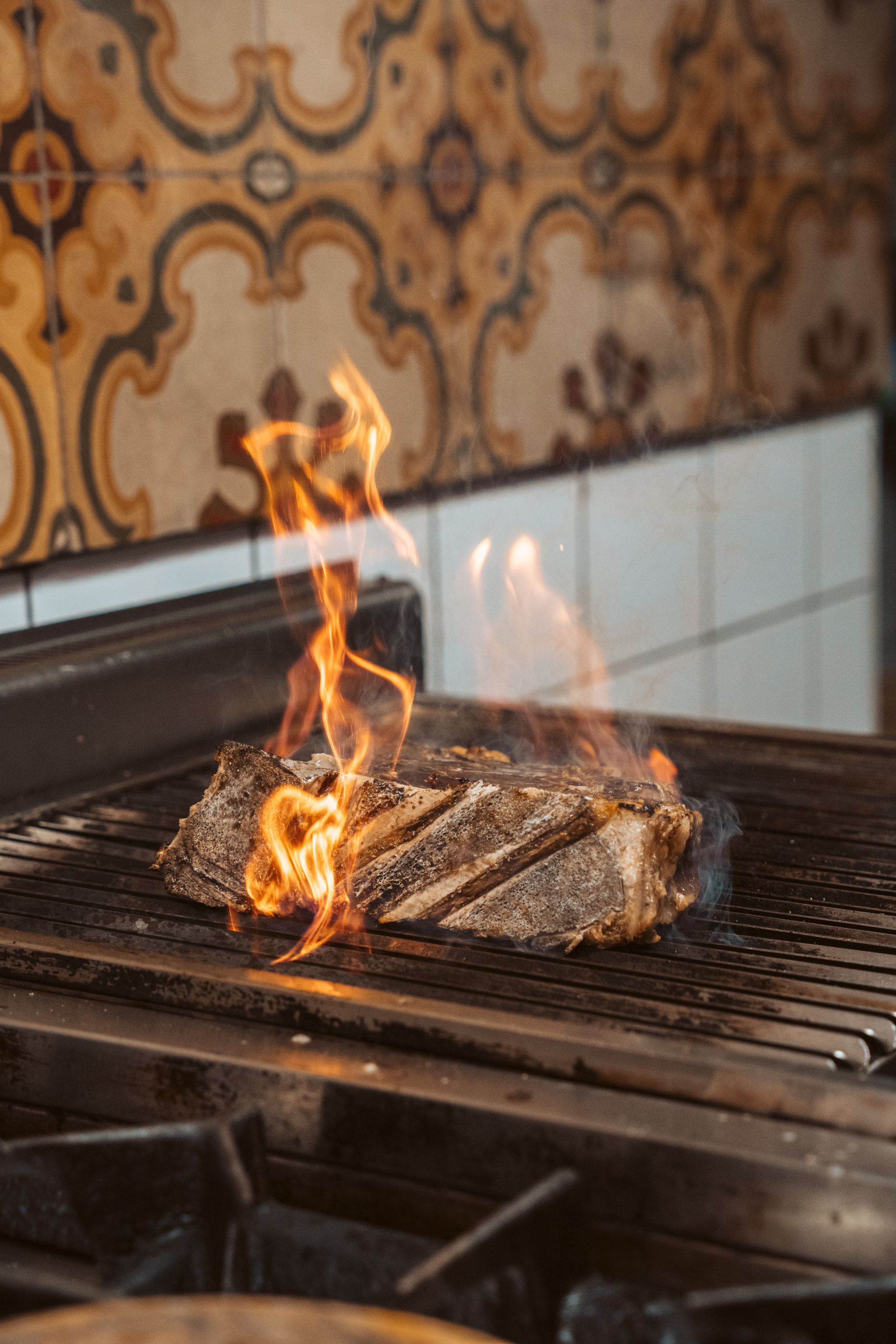 Bistecca su una griglia con fiamme, in una cucina con piastrelle decorate.