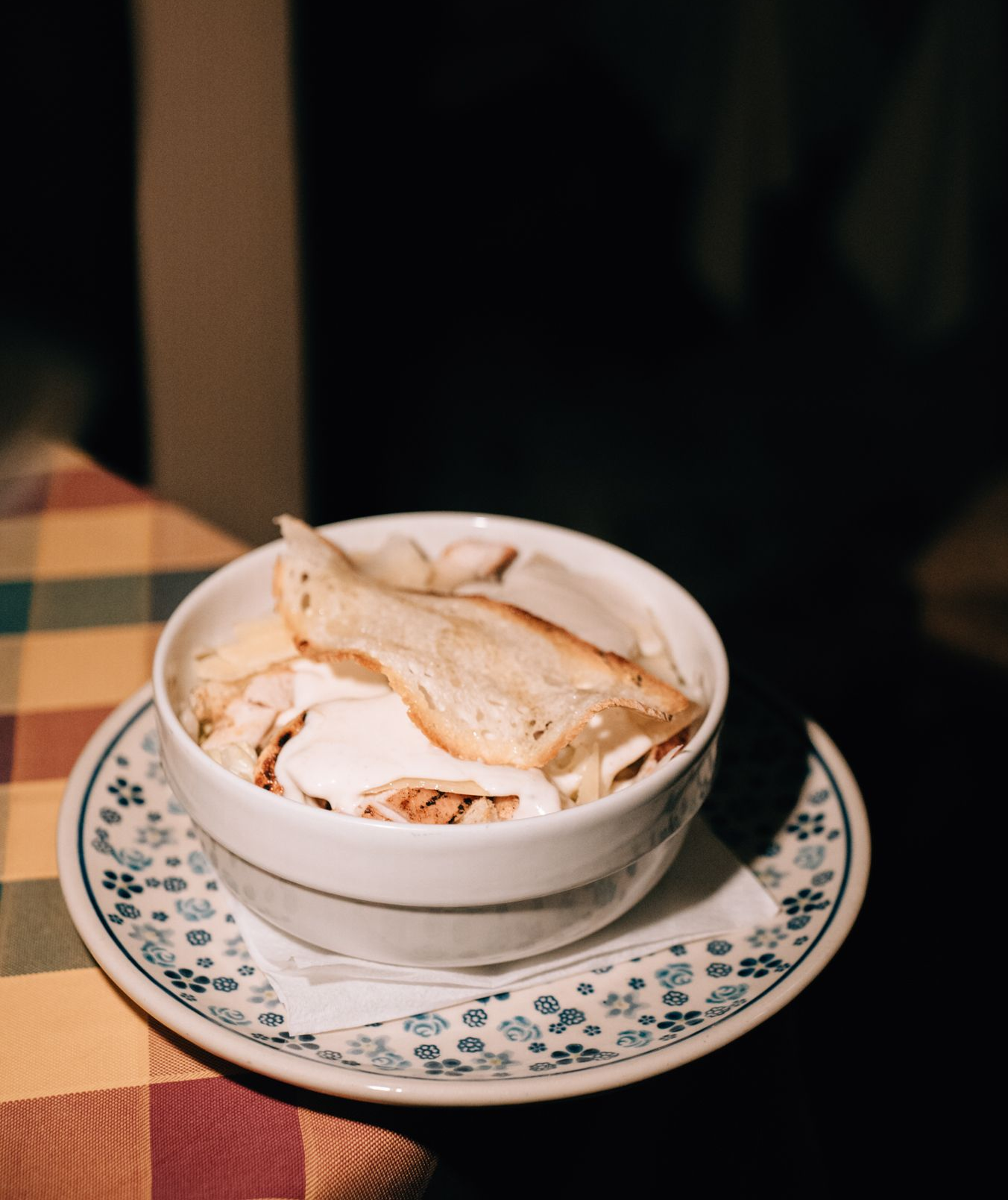 Bowl of soup with bread on a patterned saucer on a checkered tablecloth.