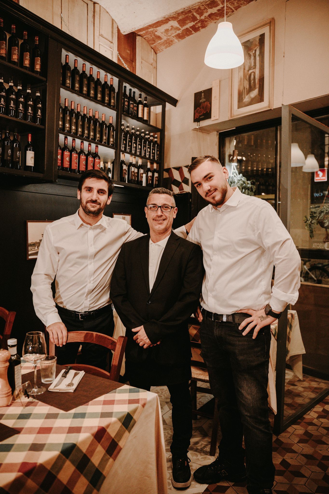 Three people pose inside a restaurant. One in a suit, two in white shirts. Wine bottles in the background.