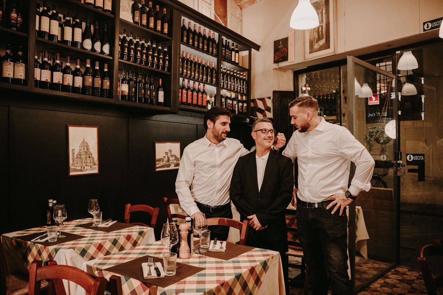 Three men in a restaurant. Two stand by a man in a blazer, near tables with checkered tablecloths and wine shelves.