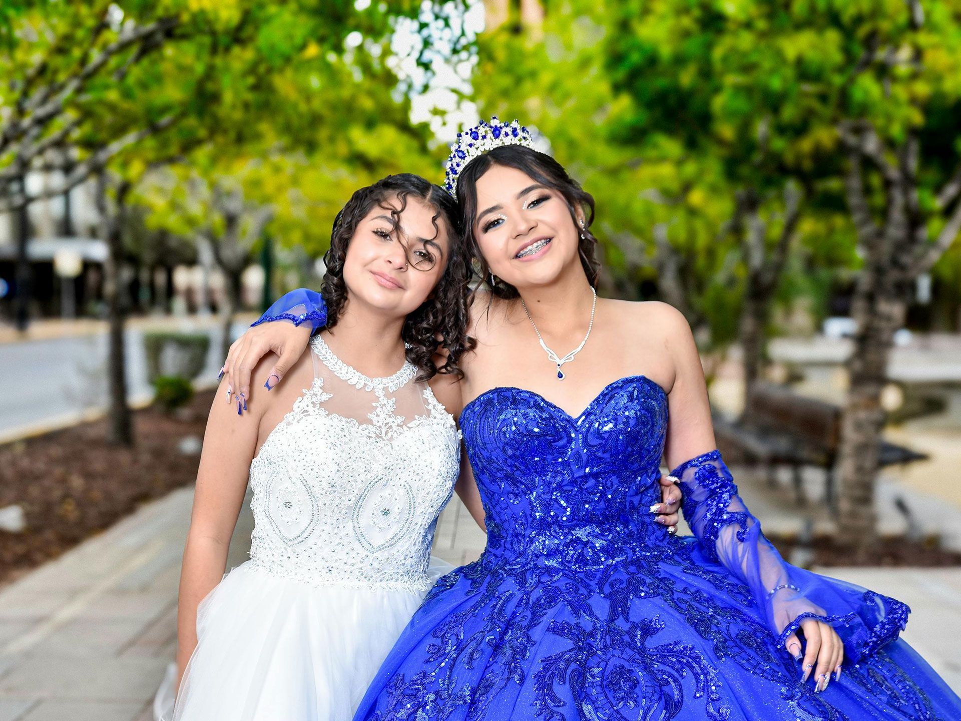 Deux personnes posent ensemble en extérieur ; l'une porte une robe blanche à perles, l'autre une robe bleue à paillettes et un diadème.