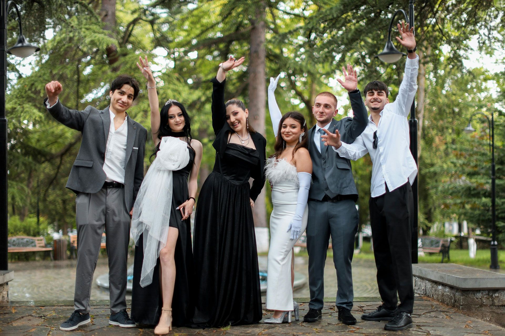 Six amis en tenue de soirée saluent la foule en posant ensemble dans un parc extérieur bordé d'arbres verdoyants.