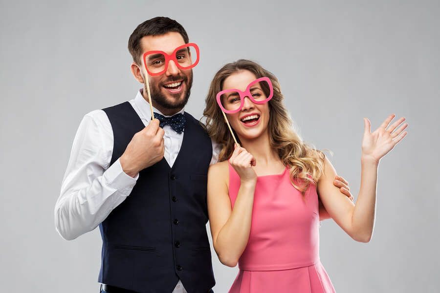 Un couple souriant pose sur un fond gris en tenant des lunettes surdimensionnées en carton rose et rouge fixées sur des bâtonnets.