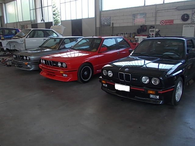 Quattro BMW M3 d'epoca in un garage. Una rossa, una nera, una grigia e una smontata.