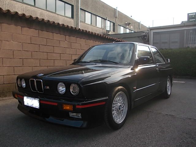 BMW E30 coupé nera parcheggiata davanti a un muro di mattoni e a un edificio. Dettagli rossi e cerchi argentati.