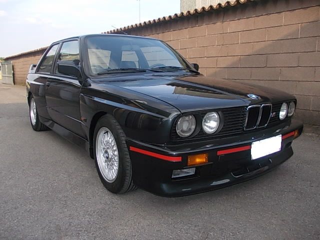BMW E30 M3 coupé nera parcheggiata sull'asfalto davanti a un muro di mattoni. Dettagli rossi sul paraurti anteriore.