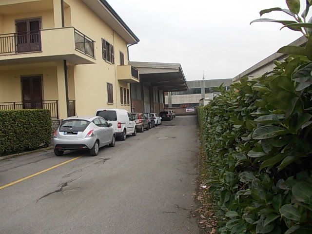Auto parcheggiate su una strada asfaltata accanto a un edificio giallo chiaro e siepi verdi sotto un cielo coperto.