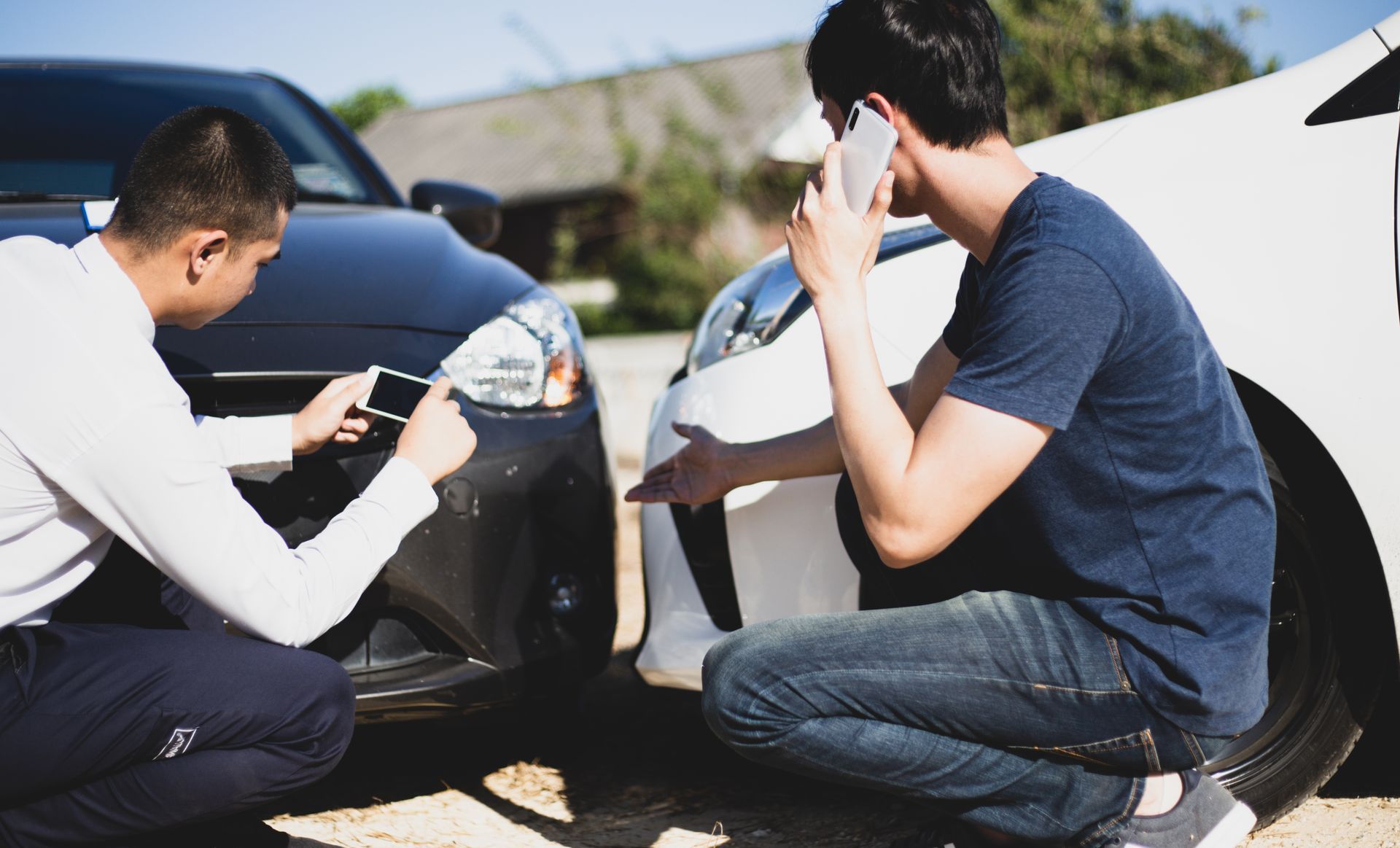Two men are kneeling next to a car and one is talking on a cell phone.