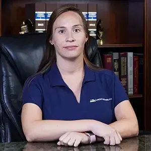 A woman in a blue shirt is sitting at a desk in front of a bookshelf.