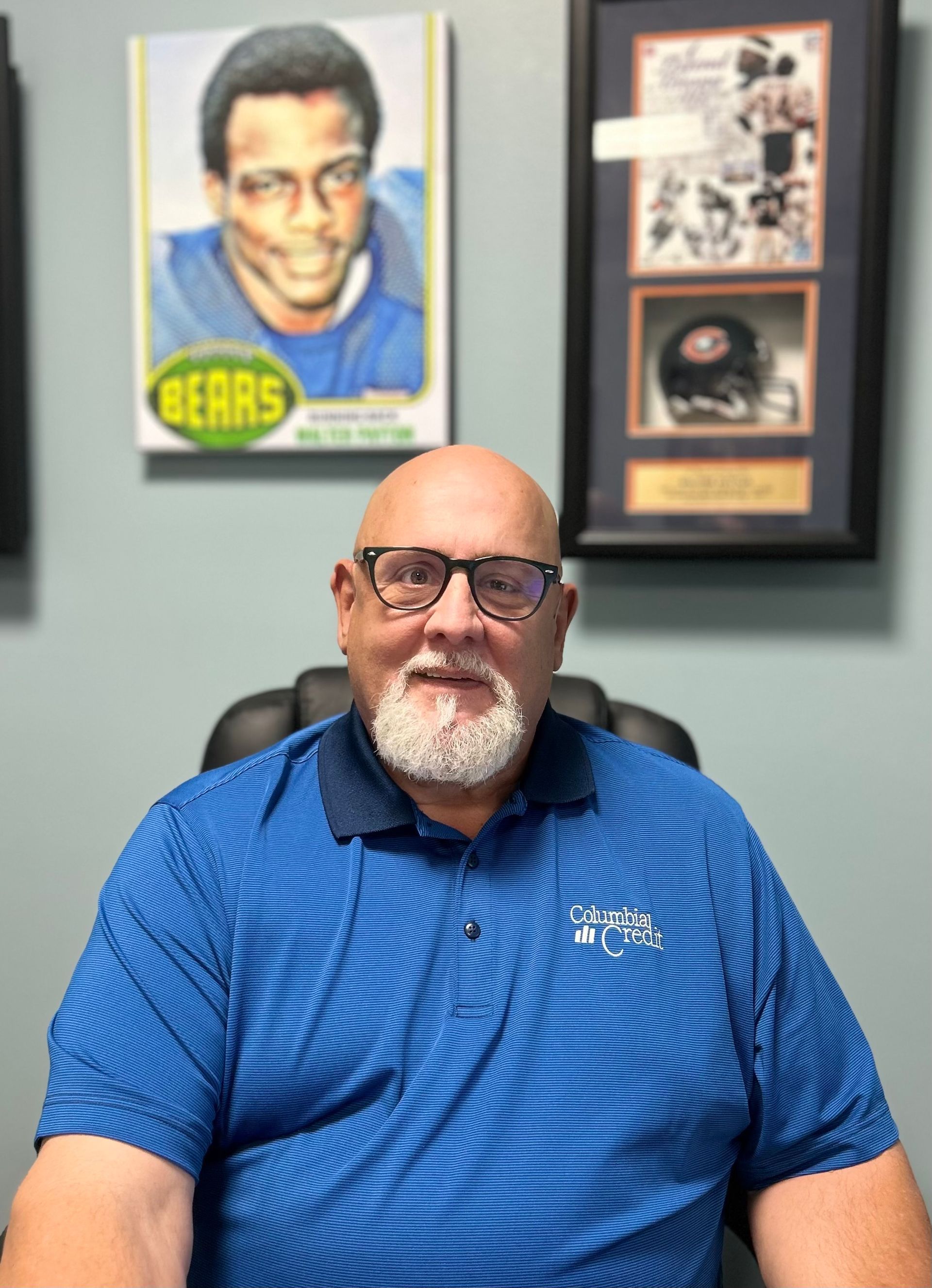 Man with glasses and beard in blue shirt, sitting at desk with Bears memorabilia.