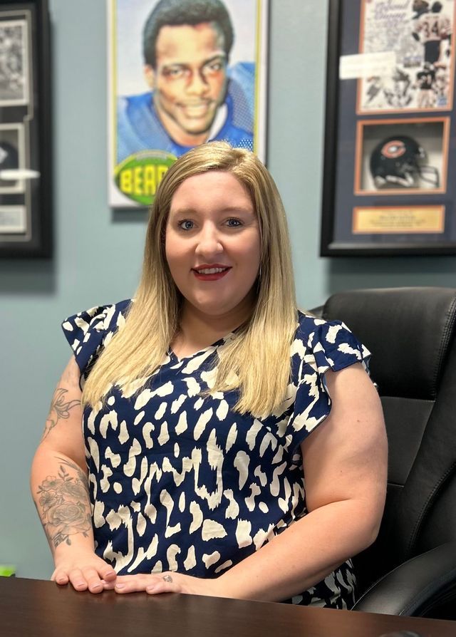 Woman with blonde hair and a patterned shirt sits at a desk, with sports memorabilia on the wall.