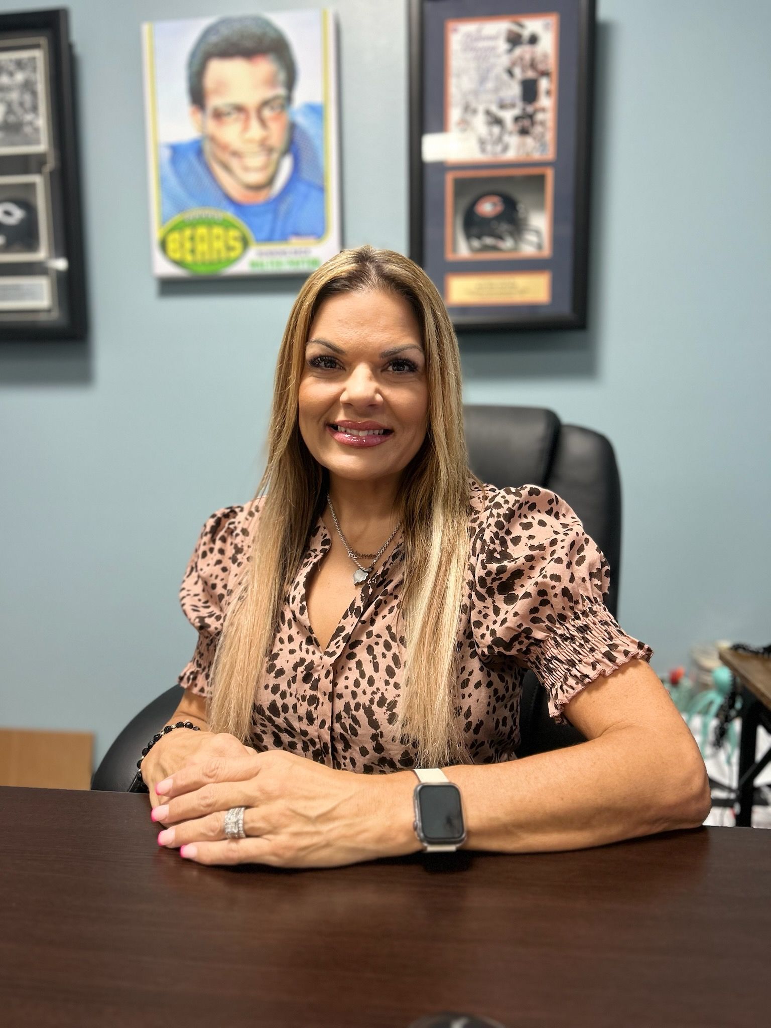 Woman with long hair, sitting at a desk, wearing a patterned blouse and jewelry, with sports memorabilia on the wall.