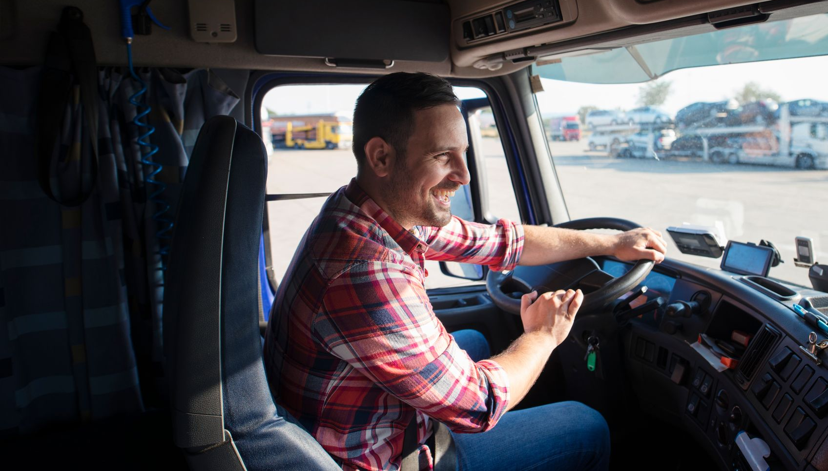 Man driving a semi-truck, smiling. Wearing red plaid shirt. Parked in a truck stop with other trucks.
