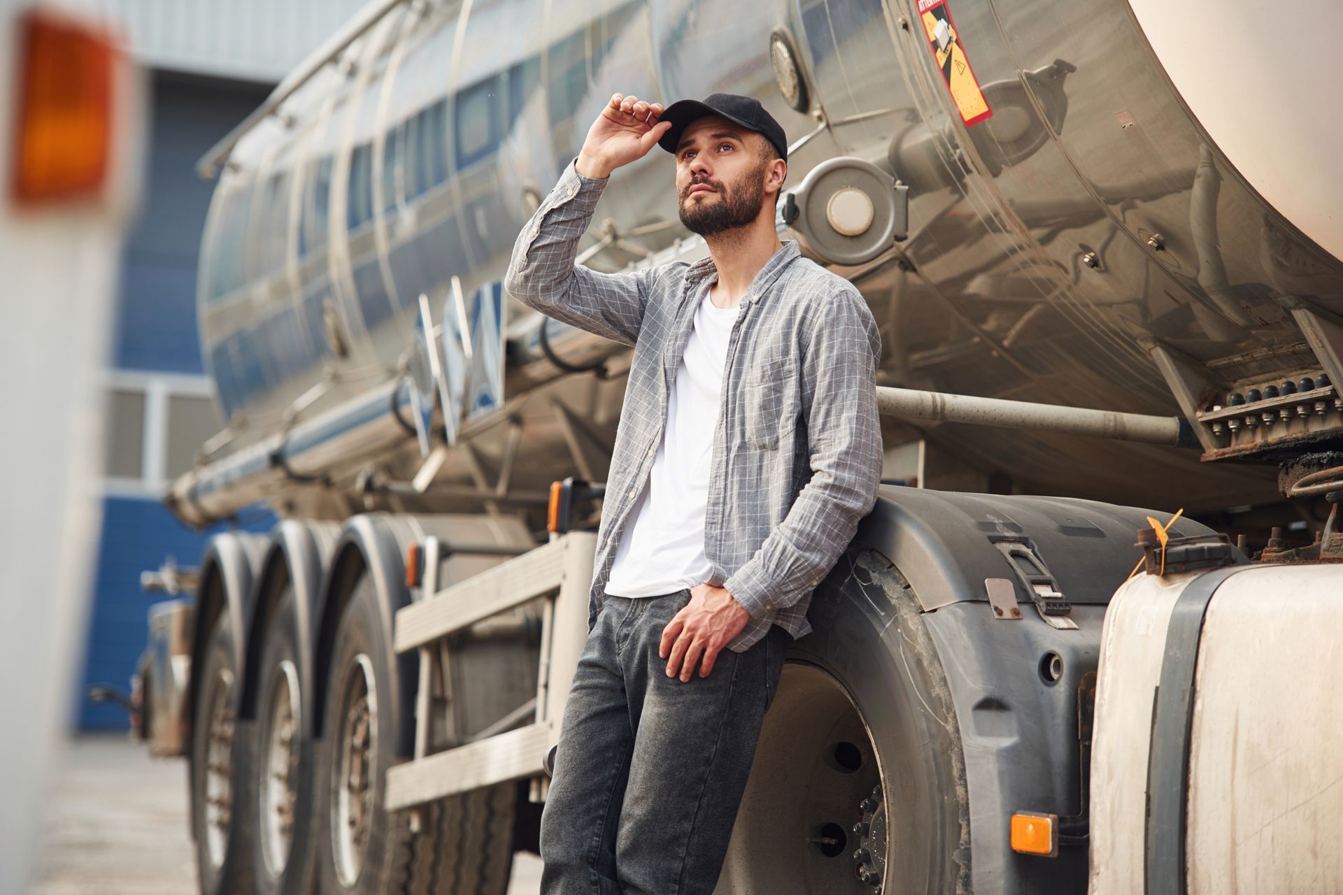 Man in a cap and shirt leans against a tanker truck, looking upward.