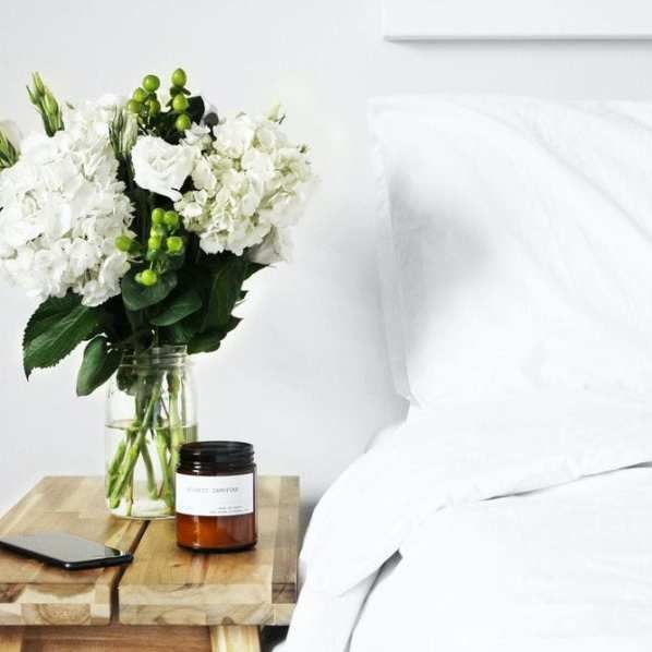 A bouquet of white flowers in a glass jar and a candle on a wooden nightstand next to a white-made bed.