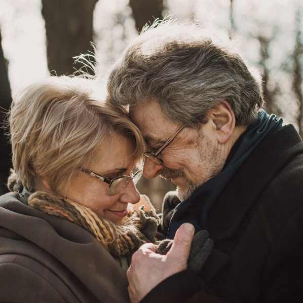Two people wearing glasses and scarves touch foreheads tenderly in a soft, outdoor setting.