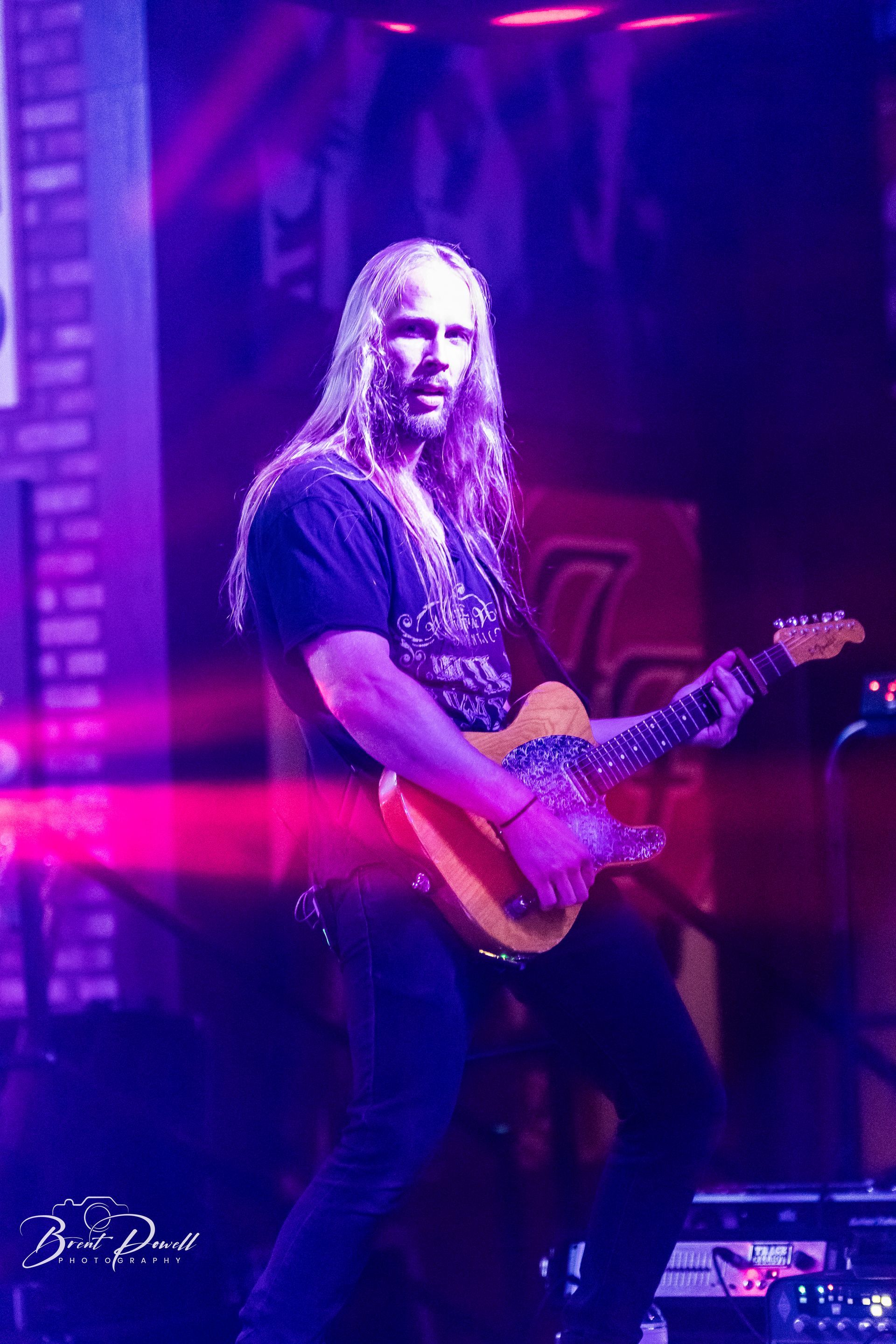 A man with long hair is playing a guitar on a stage in a dark room.