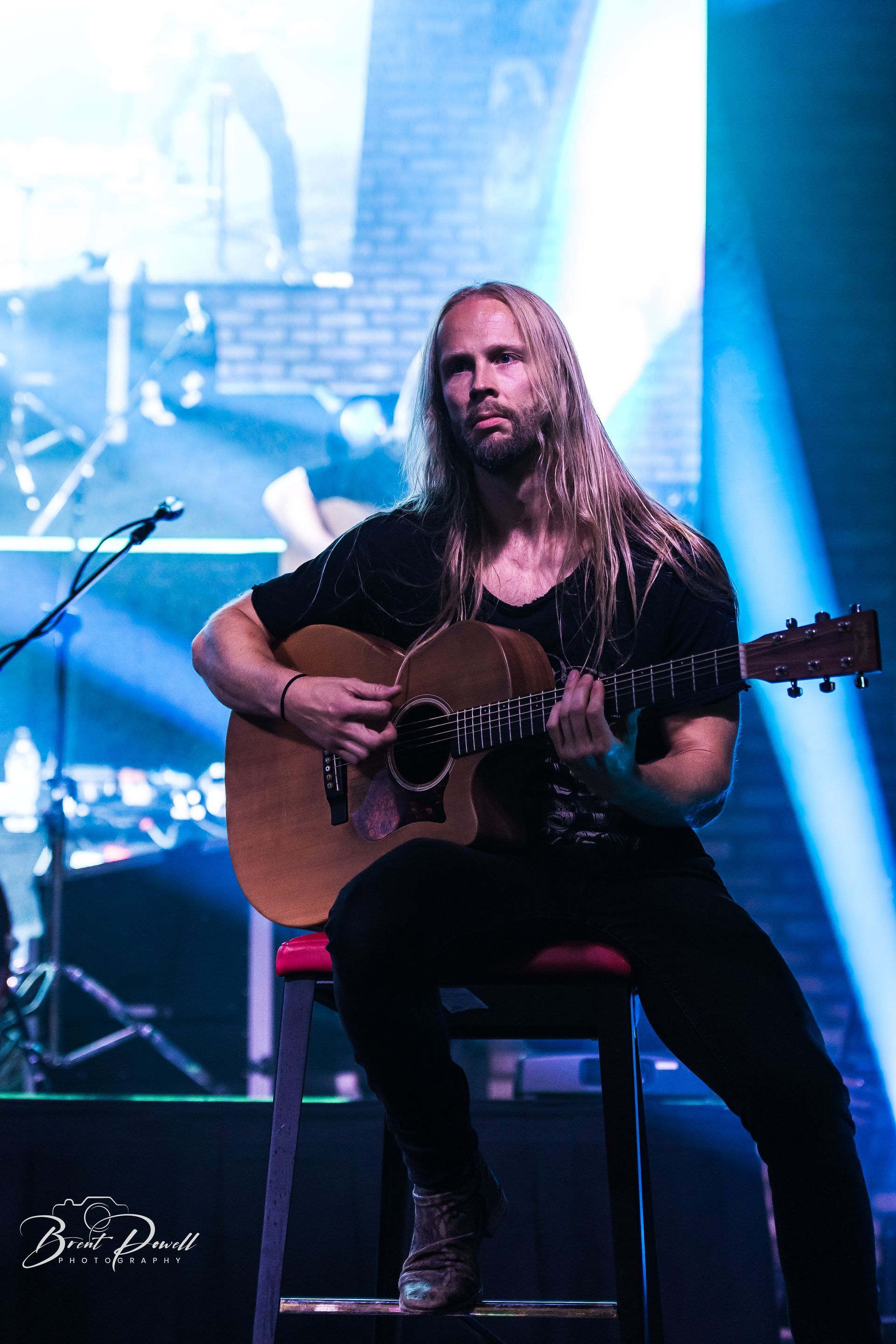 A man with long hair is sitting on a stool playing a guitar on a stage.