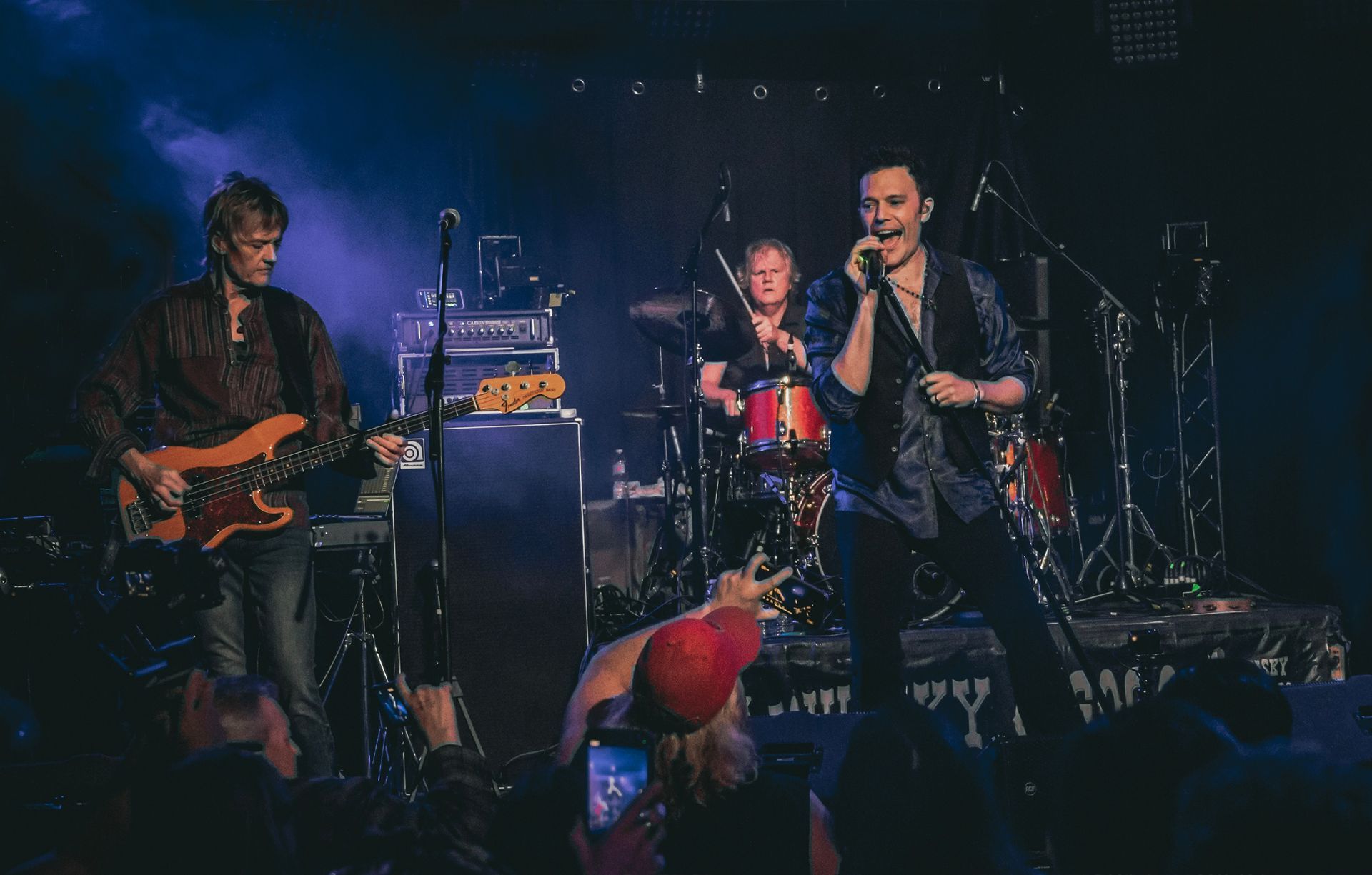 Band performing on stage: guitarist, drummer, and singer with microphone. Blue and purple stage lighting.