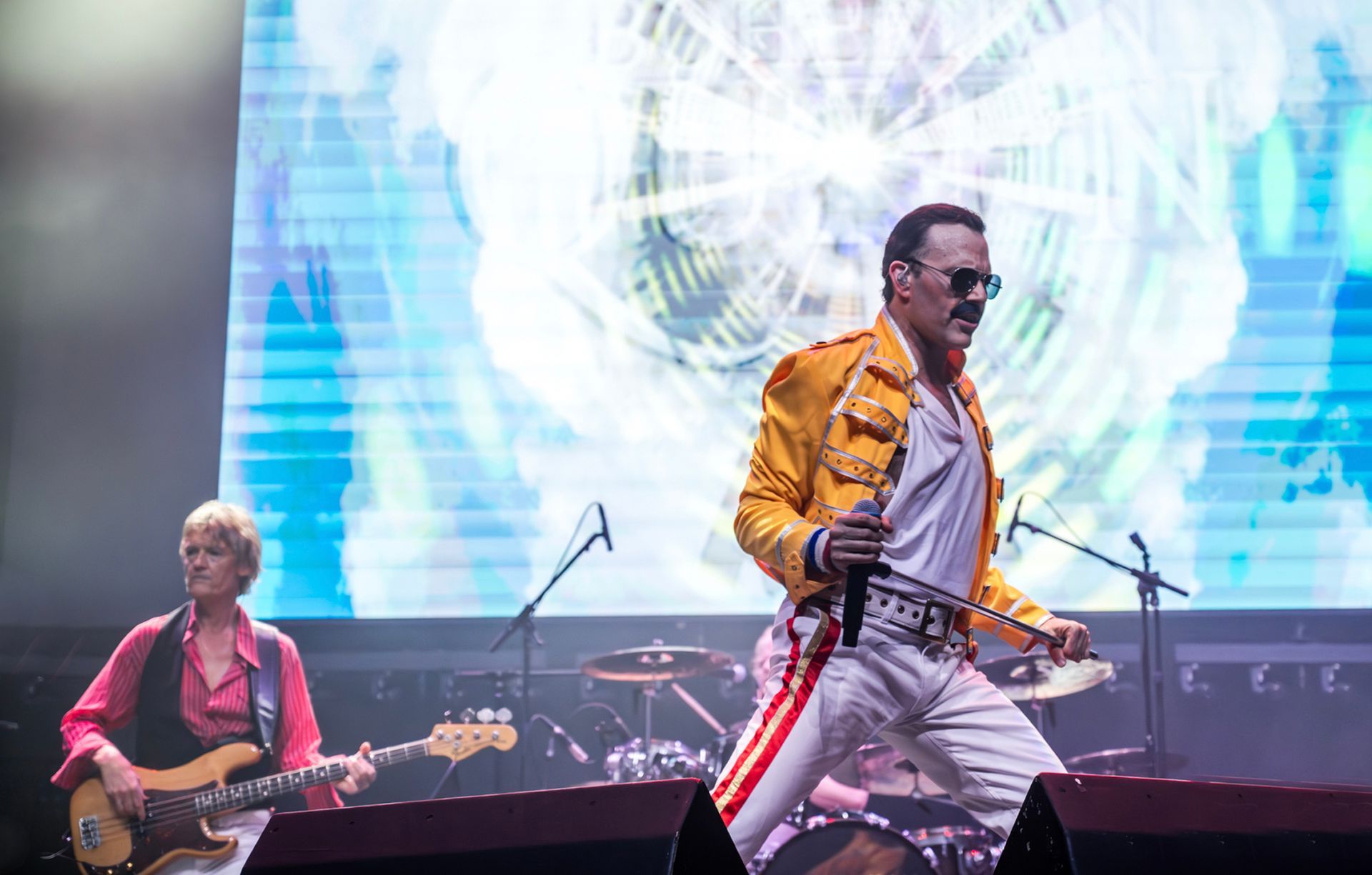 A man dressed as Freddie Mercury sings on stage with a band during a concert.