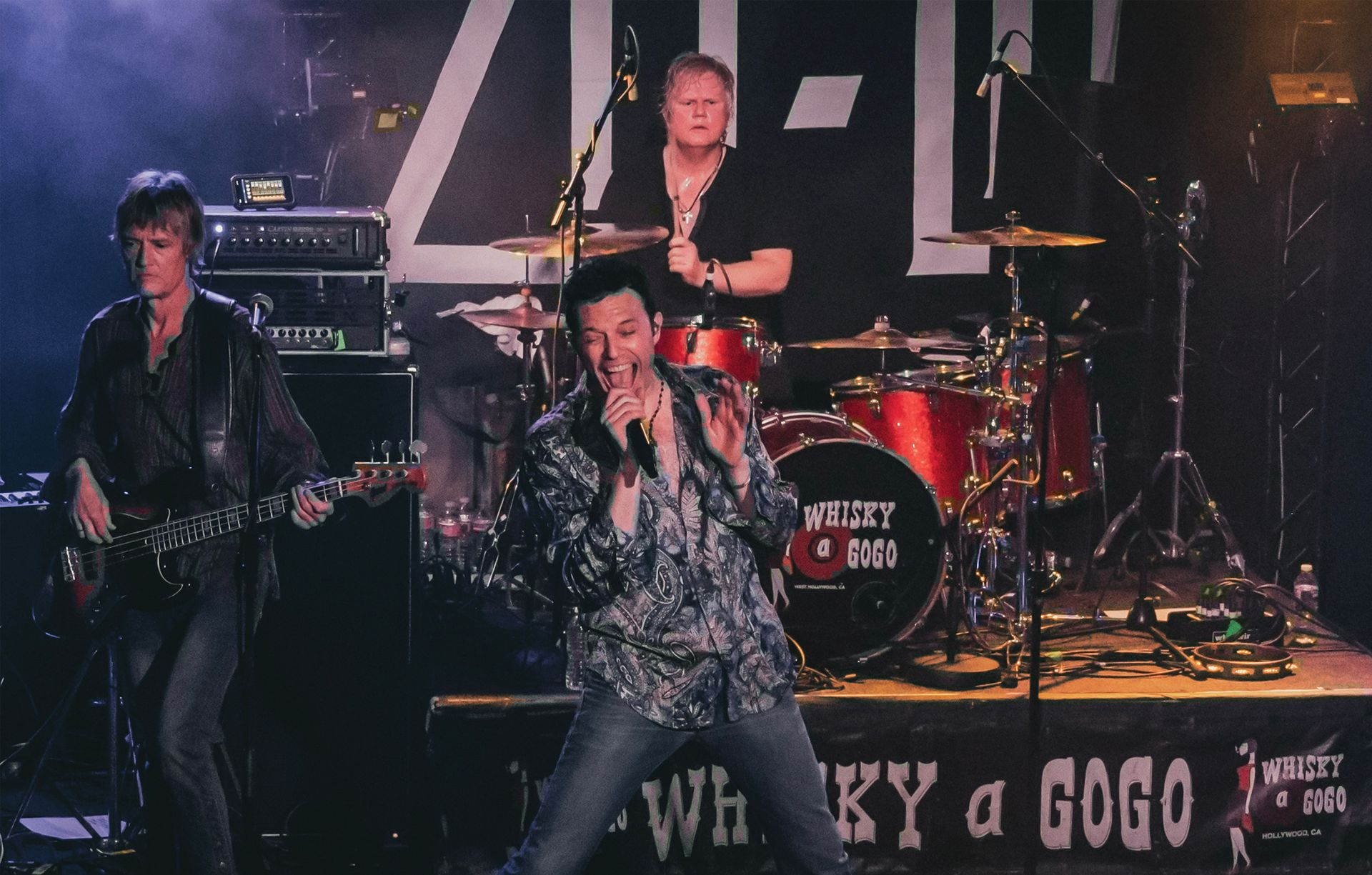 Band performing on stage at the Whisky a Go Go. Singer holds microphone, guitarist plays, drummer in background.