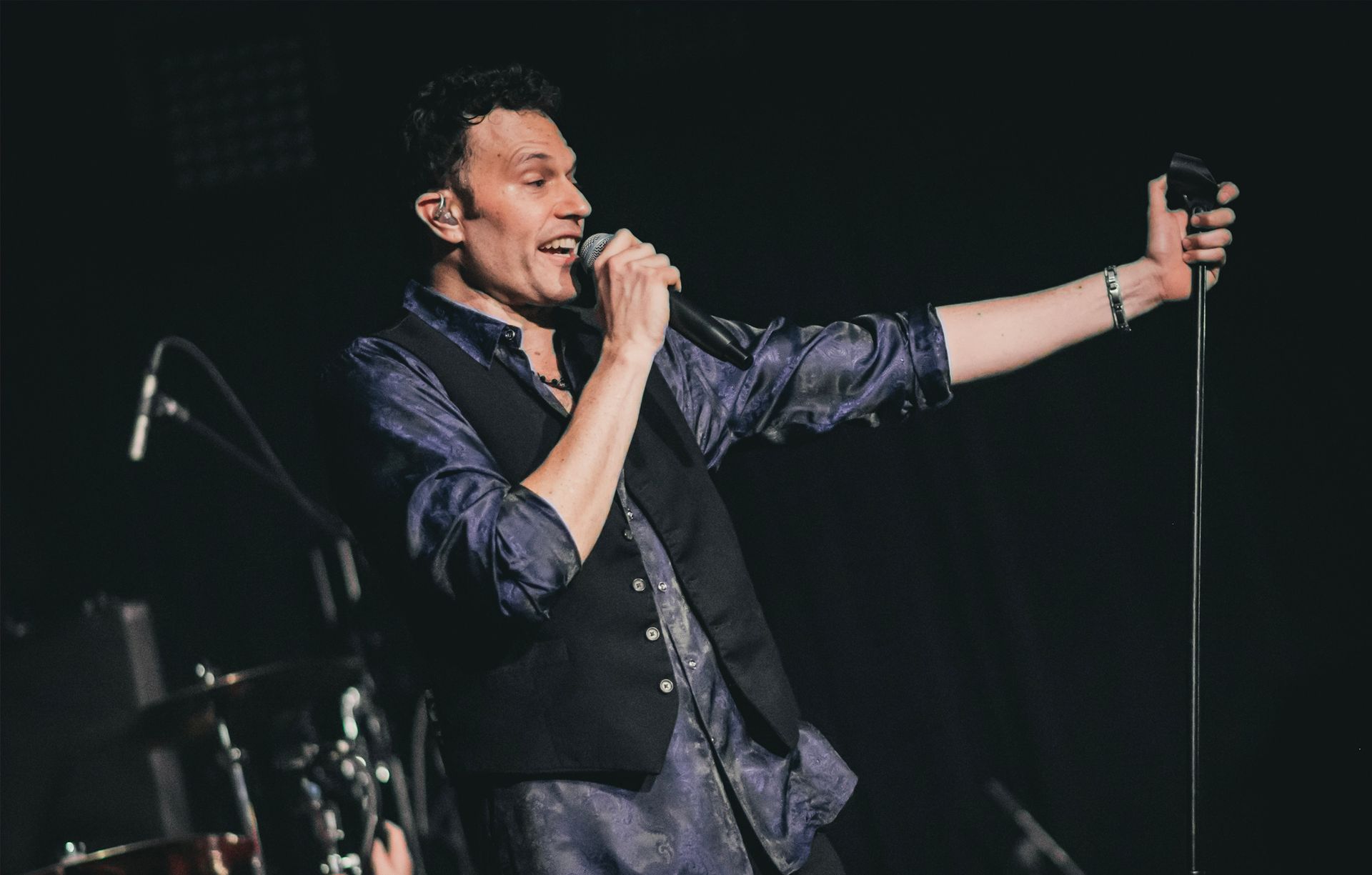 A man singing into a microphone on stage, gesturing with his right arm.
