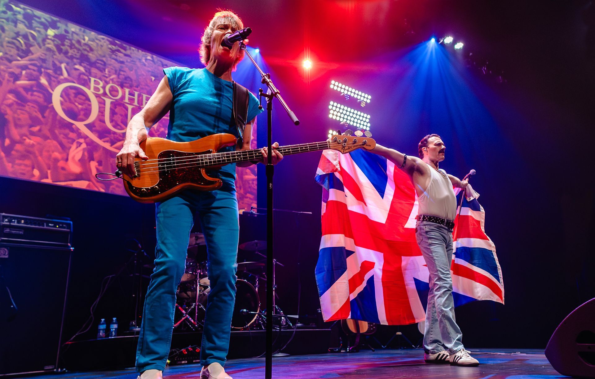 Band on stage, bassist in blue plays, singer in Freddie Mercury pose with Union Jack flag.