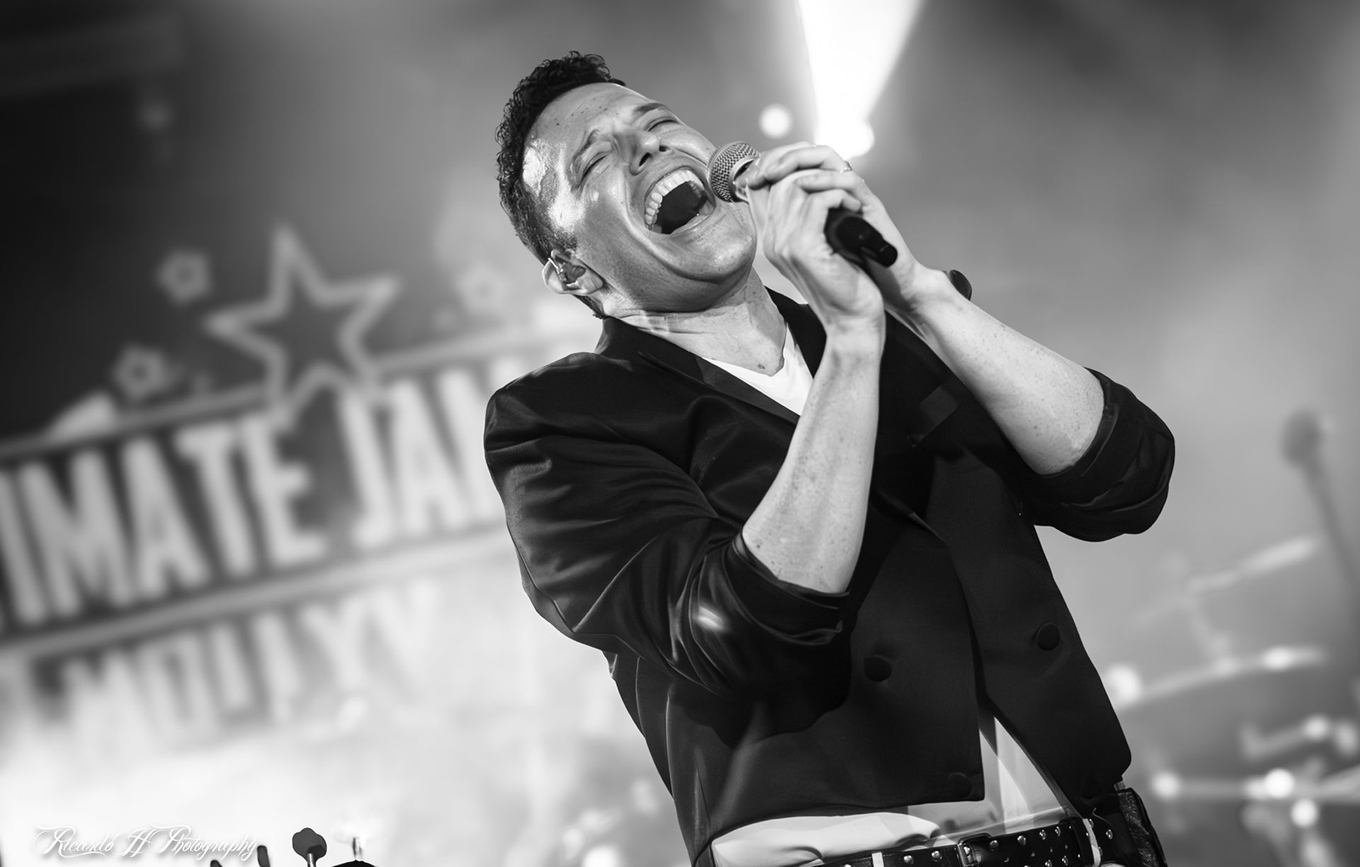 A man is singing into a microphone on a stage in a black and white photo.