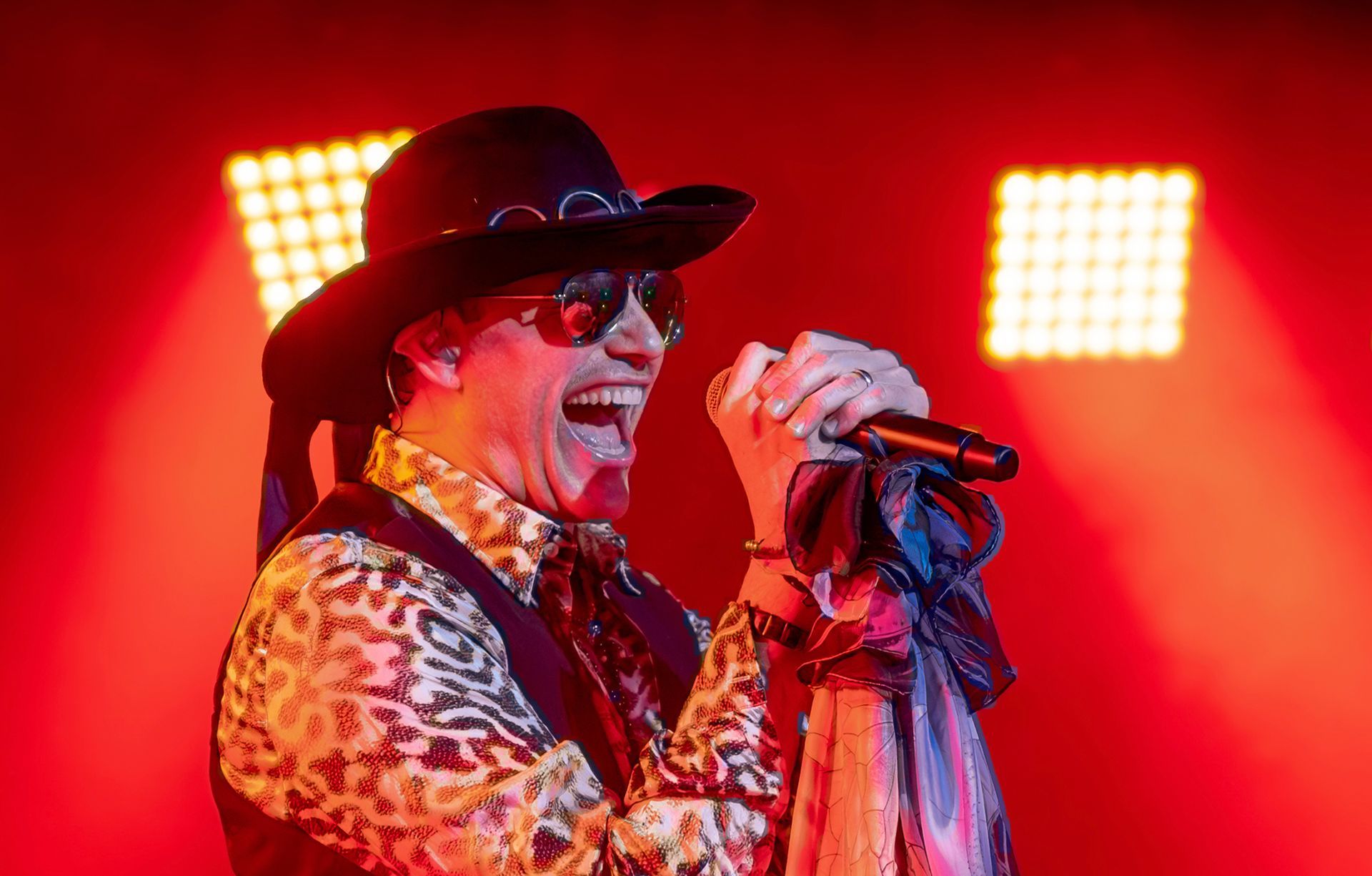 A man wearing a hat and sunglasses is singing into a microphone.